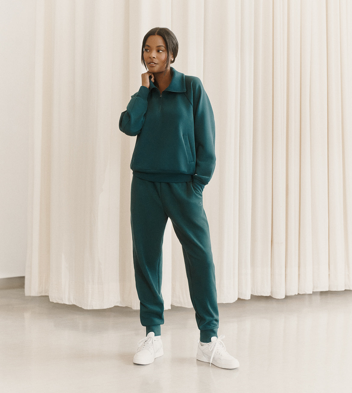 A model wearing a matching dark teal green tracksuit with a half-zip pullover sweatshirt and jogger pants, paired with white sneakers, standing in front of a cream-colored curtain backdrop.