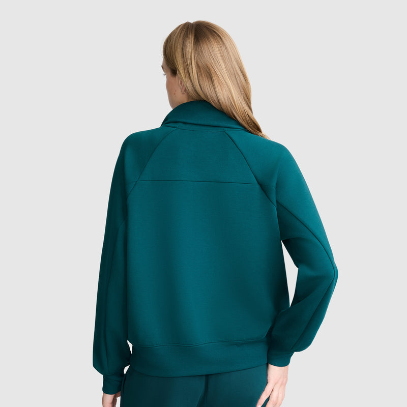 A woman with long blonde hair is seen from behind, wearing a teal Champion Quarter-Zip Pullover with matching pants, standing against a light gray background.