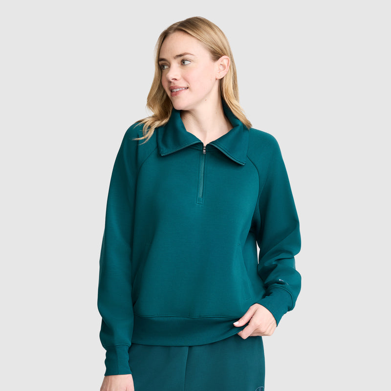 Wearing the Champion Quarter-Zip Pullover in teal stretch poly-modal, paired with matching pants, a person stands against a plain light gray background, looking to the side and smiling softly.