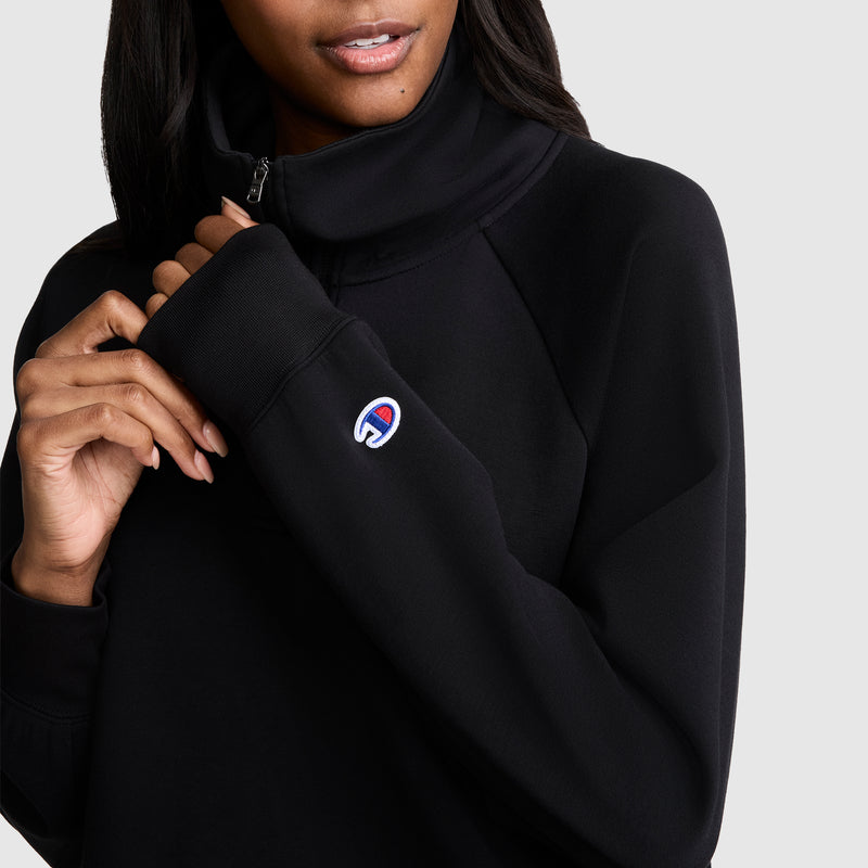 A woman wearing a Champion Quarter-Zip Pullover in black touches the collar of this stylish athletic top. The Champion logo is displayed on the sleeve, and the background is plain and light-colored.