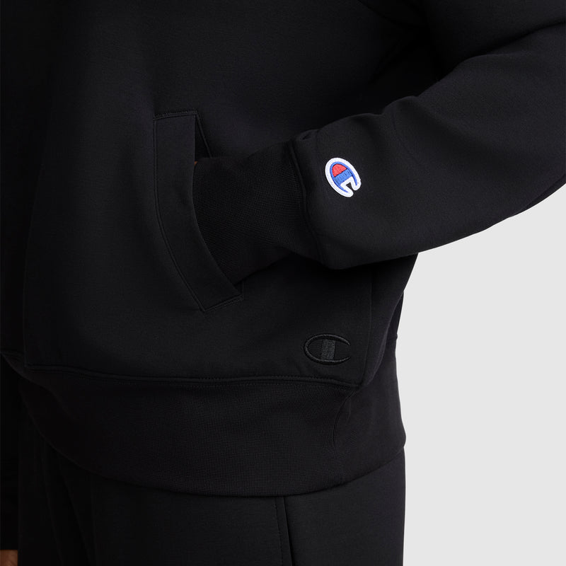 A person is wearing a Champion Quarter-Zip Pullover in black, with one hand tucked into the front pocket and the Champion logo patch visible on the sleeve. The photo is cropped to show the athletic pullover from shoulder to upper thigh.