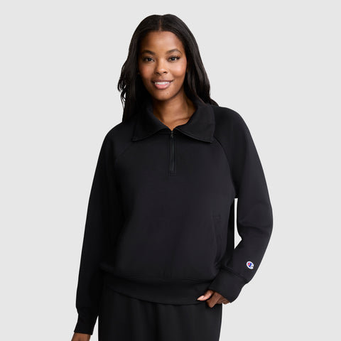 A woman stands against a plain light gray background, smiling with one hand relaxed by her side, wearing a black Champion Quarter-Zip Pullover and matching pants.