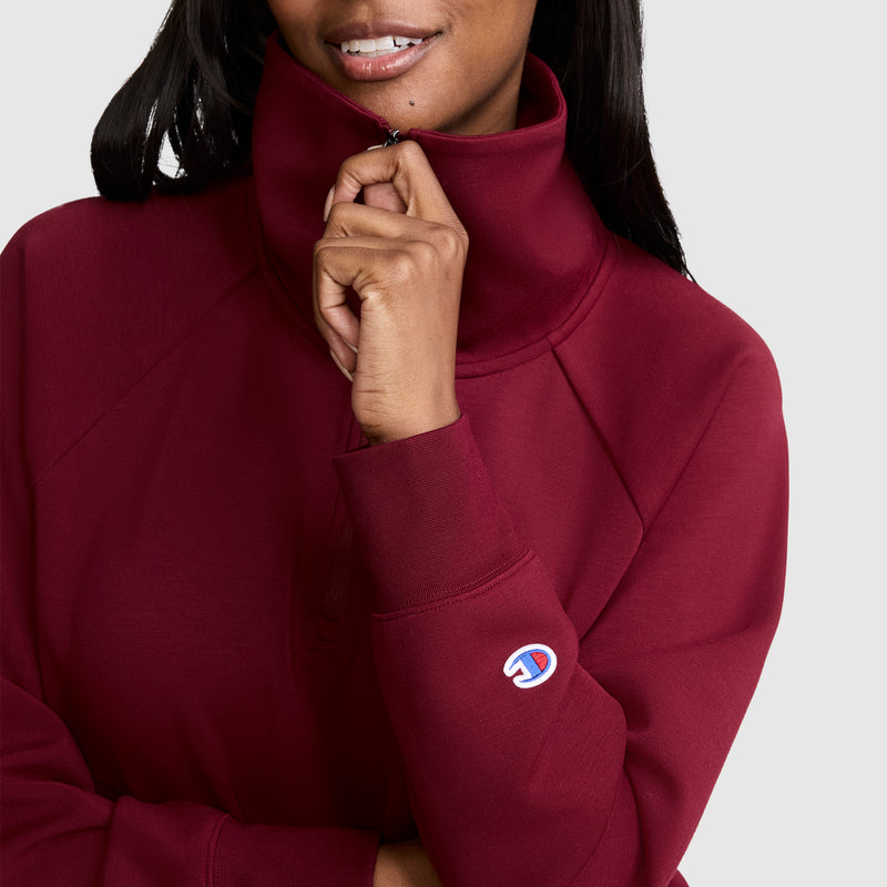A woman, smiling gently, zips up the high collar of a maroon Champion Quarter-Zip Pullover. Only her lower face and upper body are visible against a plain background.