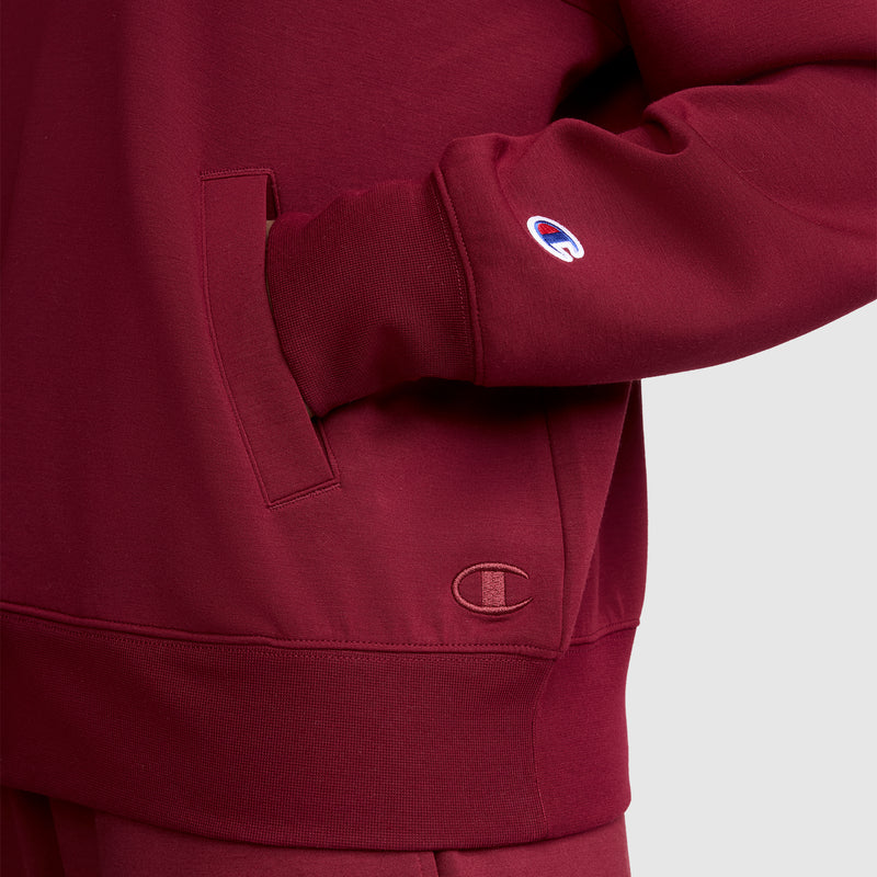 A person wearing the Champion Quarter-Zip Pullover in maroon has their hand in the pocket. This pullover, crafted from a comfortable stretch scuba blend, displays the Champion logo on the sleeve and near the hem.