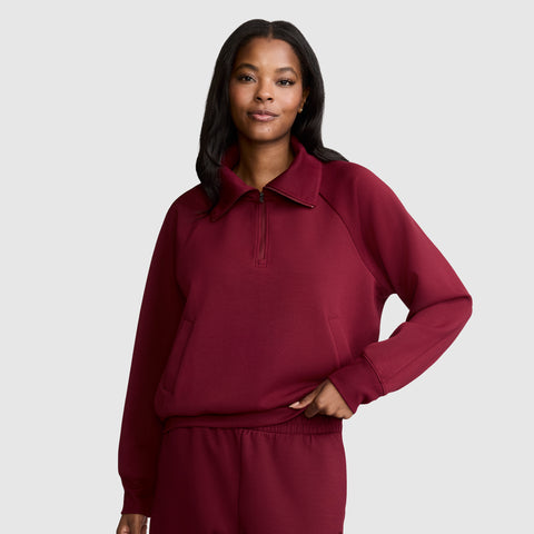 A woman stands with one hand on her hip, smiling slightly, against a plain light gray background. She is wearing a burgundy Champion Quarter-Zip Pullover and matching pants. The athletic pullover blends comfort and functionality in a stretch scuba fabric, offering an effortlessly sporty look.