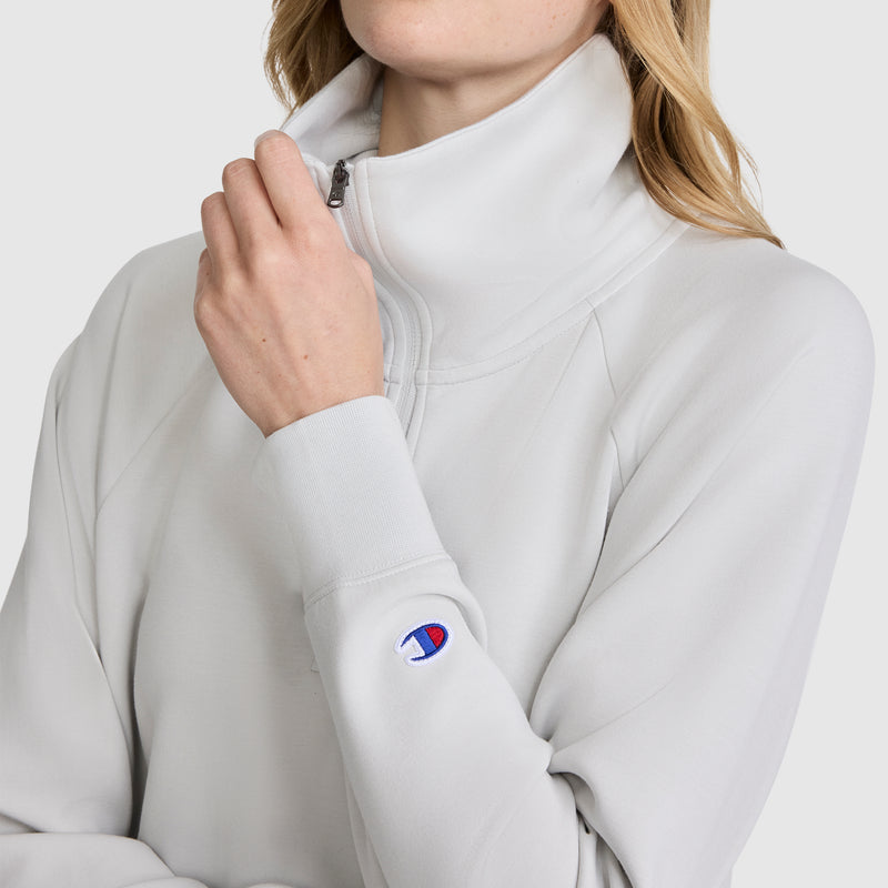A person wearing a light gray Champion Quarter-Zip Pullover made from stretch poly-modal fabric, featuring a high collar and a Champion logo patch on the sleeve, is holding the zipper with one hand. The individual’s face is partially out of frame.