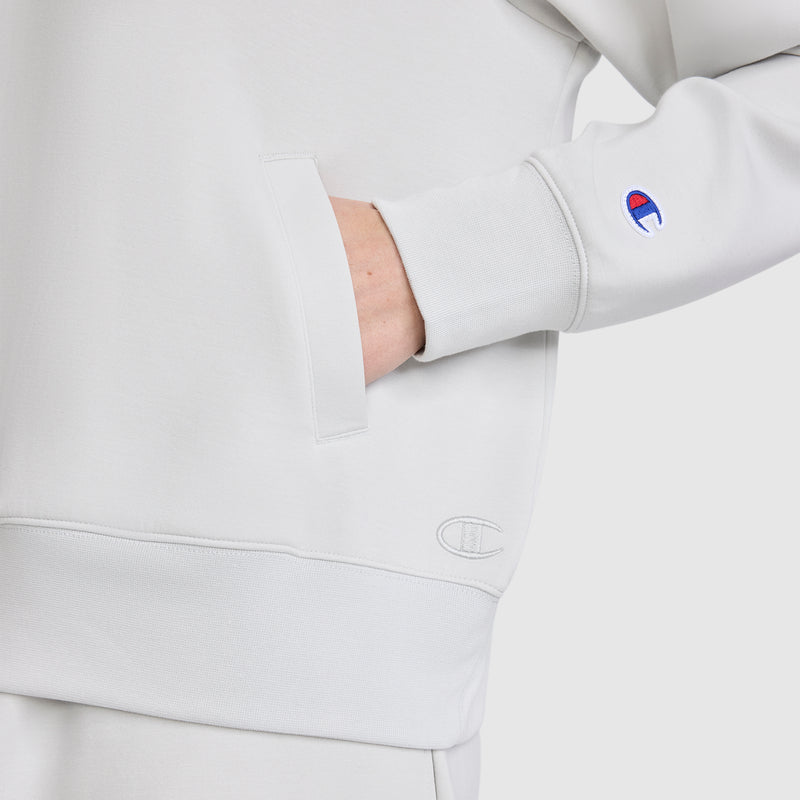 A person is wearing a light gray Champion Quarter-Zip Pullover with one hand in the front pocket. The Champion logo appears on the sleeve and near the waistband. This athletic pullover by Champion is crafted from soft, stretch poly-modal fabric for comfort and flexibility. The background is plain white.