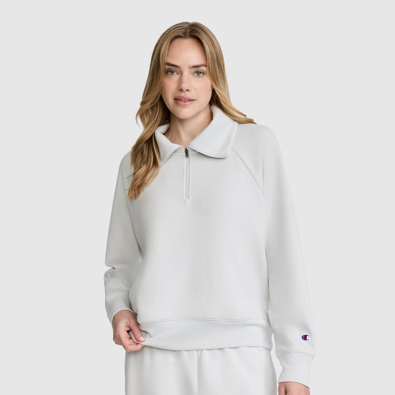 A woman stands against a plain light background, looking at the camera with a relaxed expression. She’s wearing a light gray Champion Quarter-Zip Pullover and matching stretch poly-modal pants.