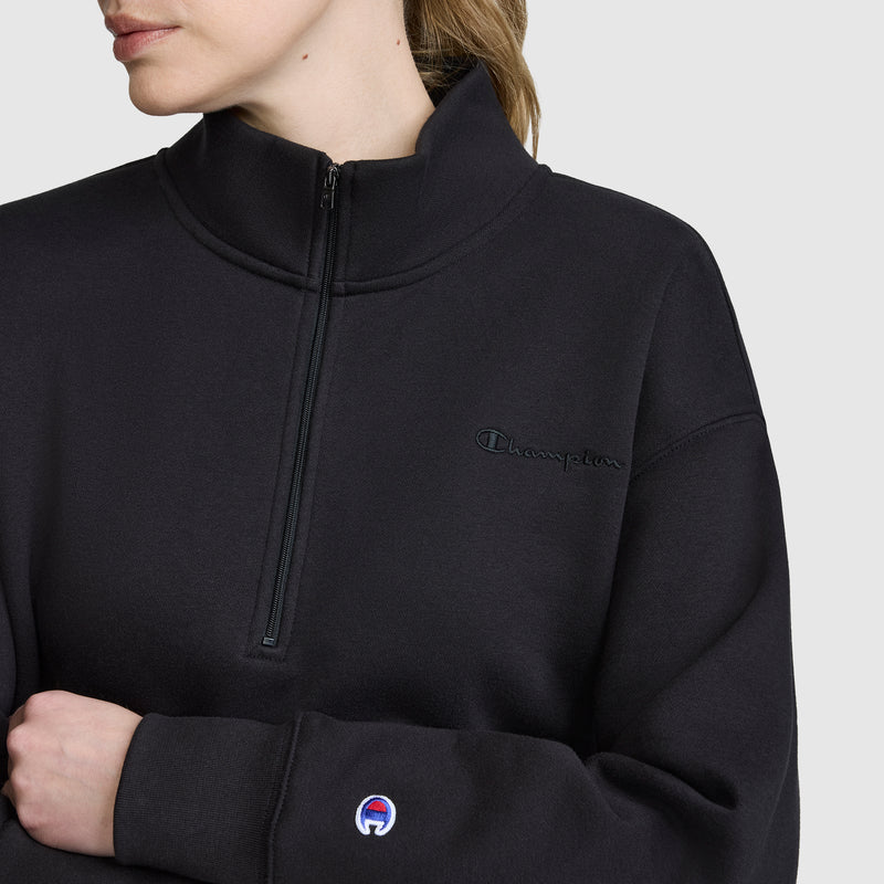 A woman wears the Champion Fleece Mockneck Quarter-Zip sweatshirt in black, featuring the logo on the chest and cuff, styled for a sleek sport-inspired look against a plain light background.
