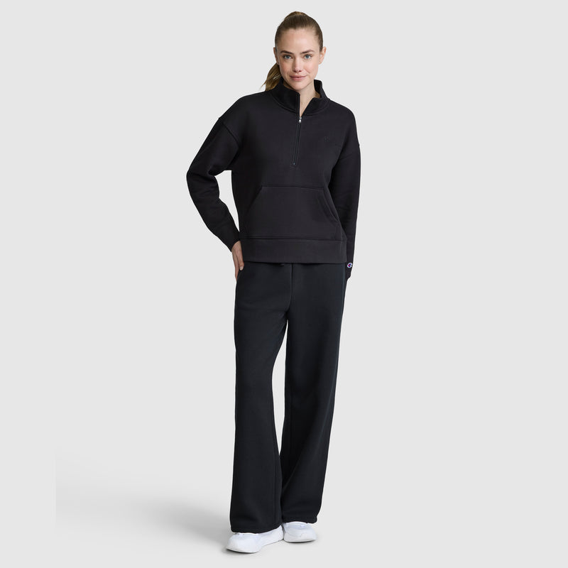 A person stands facing forward, wearing the Champion Fleece Mockneck Quarter-Zip with matching black wide-leg pants and white sneakers, showcasing a modern sport style against a plain light gray background.