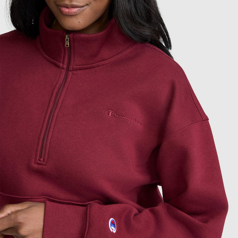 A woman, shown from the shoulders down, wears a maroon Champion Fleece Mockneck Quarter-Zip sweatshirt with the brand’s embroidered logo on the chest and sleeve, capturing an effortless sporty style.