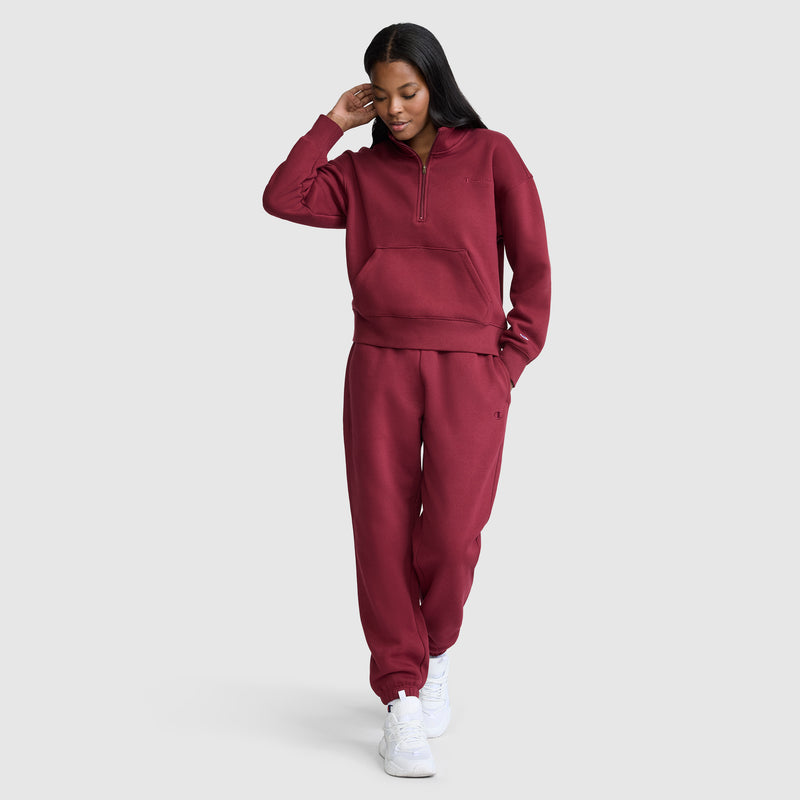 A woman wearing a maroon Champion Fleece Mockneck Quarter-Zip and white sneakers stands against a light gray background, looking down and touching her hair with one hand while the other is in her pocket, effortlessly embodying sport style.