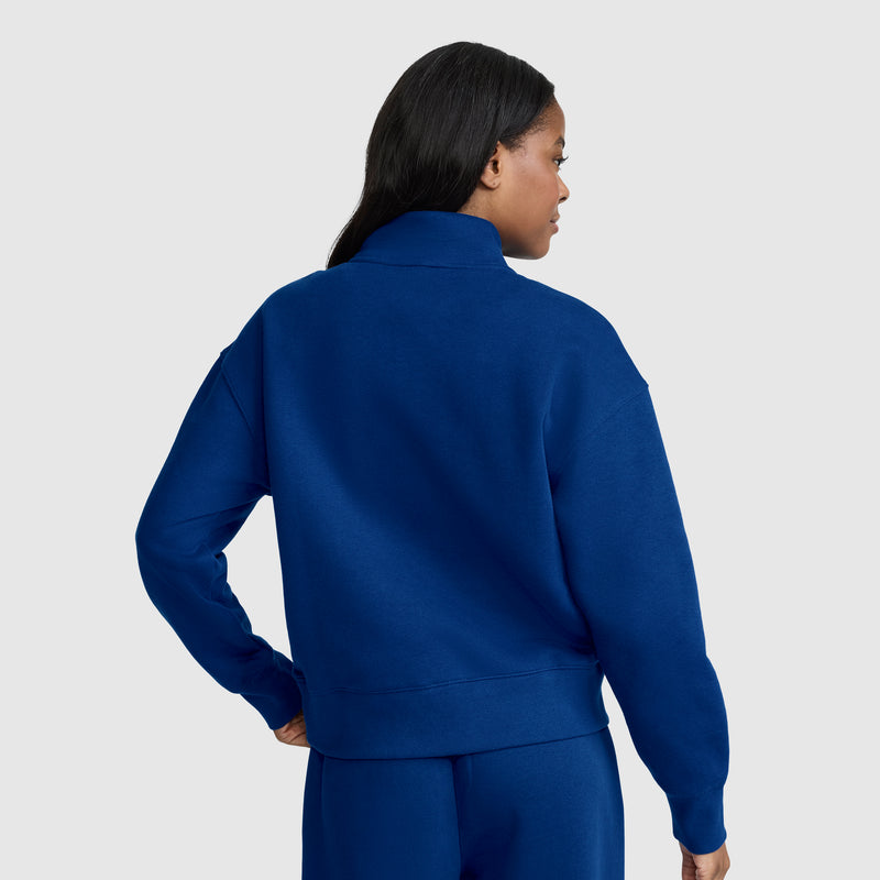 A woman with long dark hair is seen from behind against a plain light gray background, wearing a royal blue Champion Fleece Mockneck Quarter-Zip sweatshirt with matching pants.