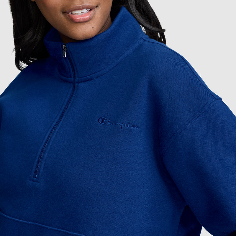 A woman wearing a royal blue Champion Fleece Mockneck Quarter-Zip, featuring the iconic Champion logo on the chest, stands against a plain light background and smiles softly, embodying timeless sport style.