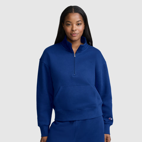 A woman stands against a plain light gray background wearing a royal blue Champion Fleece Mockneck Quarter-Zip sweatshirt with a front pocket and matching pants.