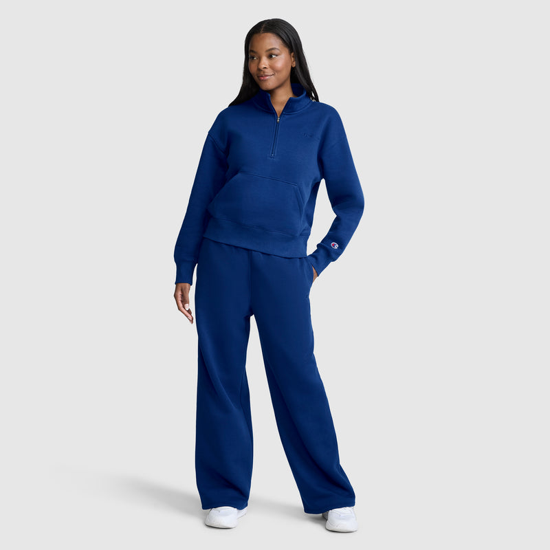 A woman stands smiling, wearing the Champion Fleece Mockneck Quarter-Zip paired with matching wide-leg sweatpants and white sneakers. Featuring a comfortable mock neck quarter-zip design, the outfit showcases a casual, heritage sport style as she poses against a plain light gray background.
