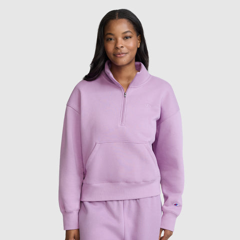 A woman stands facing forward, smiling slightly, against a plain light gray background, dressed in a light purple Champion Fleece Mockneck Quarter-Zip and matching pants that showcase a casual sport style.