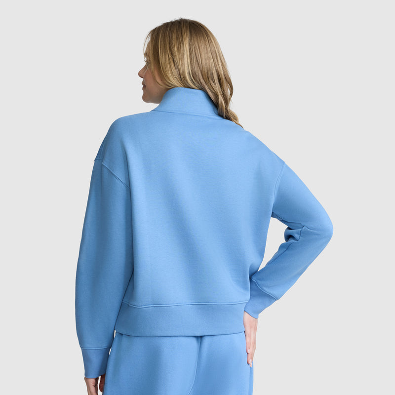 A woman with long blonde hair stands with her back to the camera, left hand on her hip, modeling the Champion Fleece Mockneck Quarter-Zip and matching pants in a classic sport style against a light gray background.