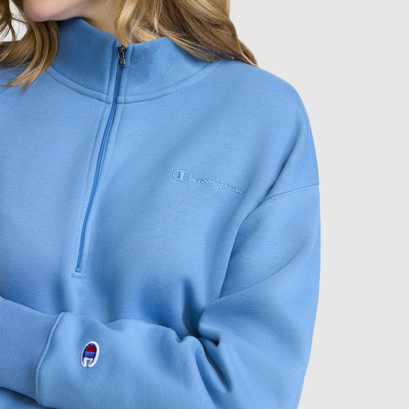 Clad in a light blue Champion Fleece Mockneck Quarter-Zip featuring embroidered logos on the chest and sleeve, this look effortlessly showcases sporty style against a simple light backdrop.