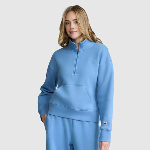A blonde-haired woman models the Champion Fleece Mockneck Quarter-Zip in light blue with coordinating sweatpants, standing with one hand in her pocket against a plain light background for a relaxed sporty look.