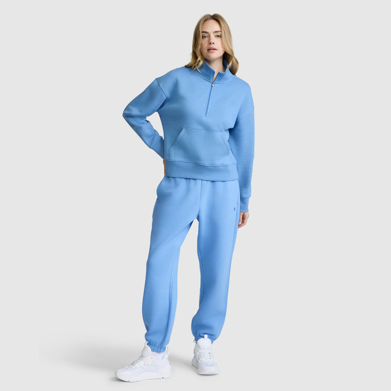 A person stands facing forward, hands relaxed, dressed in a matching light blue tracksuit featuring the Champion Fleece Mockneck Quarter-Zip sweatshirt and jogger pants. Embracing a classic sporty look, they wear white sneakers and have shoulder-length blonde hair. The background is plain light gray.