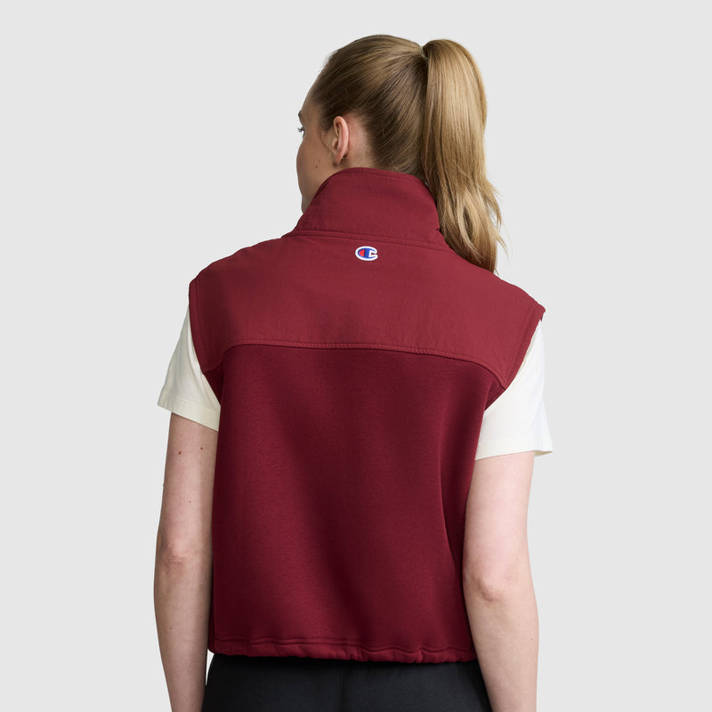 A person with blond hair in a ponytail is wearing a white short-sleeve shirt and the maroon Champion Training Vest, which features a small logo on the back, standing against a plain gray background.