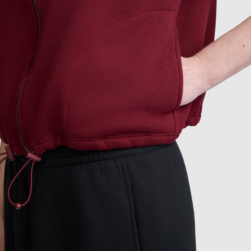 A person wearing the Champion Training Vest in maroon, styled as an athletic layering piece with their hand in the pocket and paired with black pants, shown from the waist to mid-arm against a plain light background.