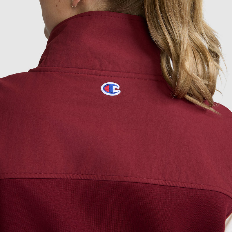 A person with light brown hair is wearing the Champion Training Vest, a red jacket with a raised collar designed for athletic layering. The Champion logo is embroidered on the back near the neck. The background is plain and light gray.