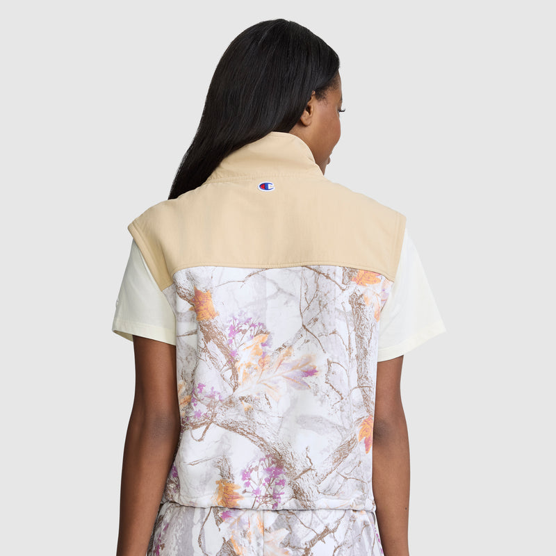 A person with long dark hair, seen from behind against a plain background, is wearing the Champion Training Vest, Allover Print—a short-sleeve top featuring a beige upper section and a light tree-branch pattern reminiscent of camouflage.