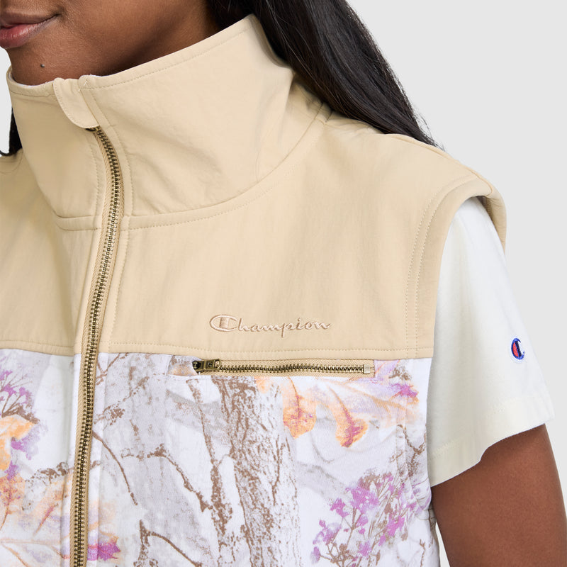 A woman wears the Champion Training Vest in an allover floral and beige print, featuring a sleeveless design, high collar, and zippered chest pocket over a white T-shirt. The Champion logo appears on both the vest and the T-shirt sleeve, combining athletic style with functional flair.