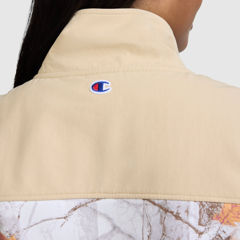 Here is a revised version of the sentence using your product data: Close-up of the back of the Champion Training Vest, Allover Print in beige, featuring the Champion logo stitched near the collar. The lower part of this women’s vest showcases an allover white and brown marble-like pattern, blending sporty appeal with unique design details.