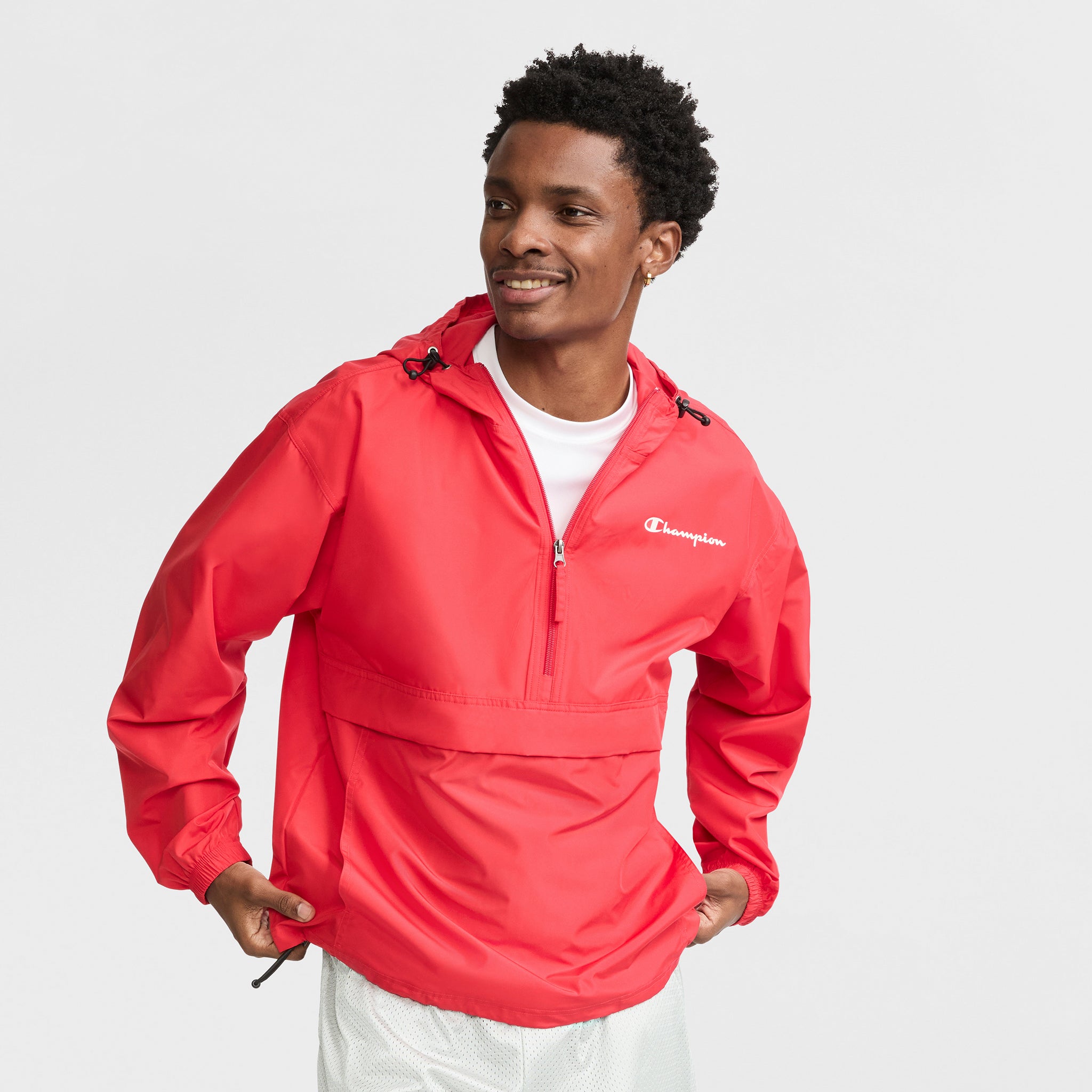 A smiling man wears the Champion Stadium Packable Jacket, Script Logo—a bright red, ultra-lightweight unisex windbreaker with front zipper and hood—over a white shirt and light shorts, standing against a plain white background.