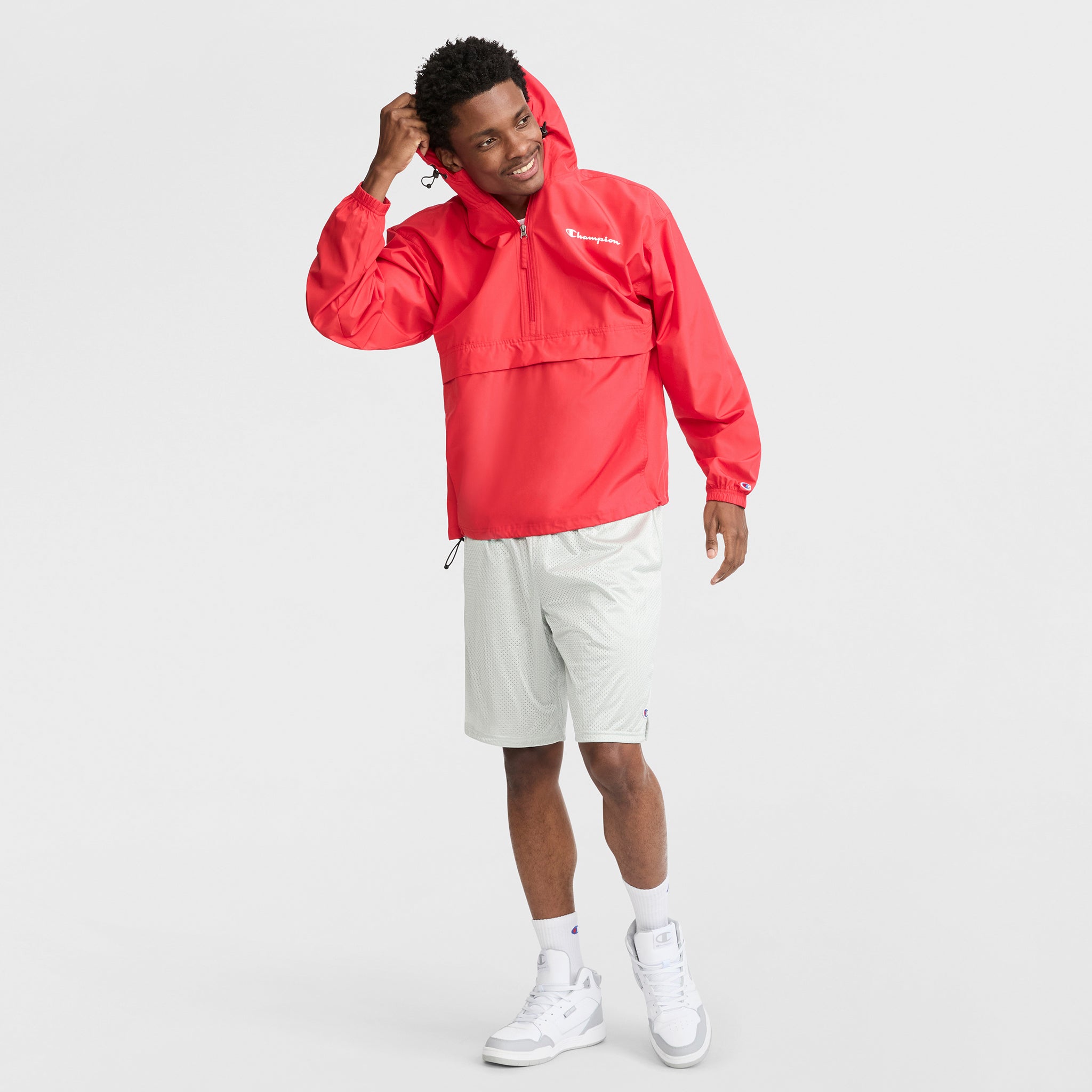 A man in a Champion Stadium Packable Jacket with Script Logo (bright red), light gray shorts, and white high-top sneakers stands smiling with one hand touching his hair against a plain white background.