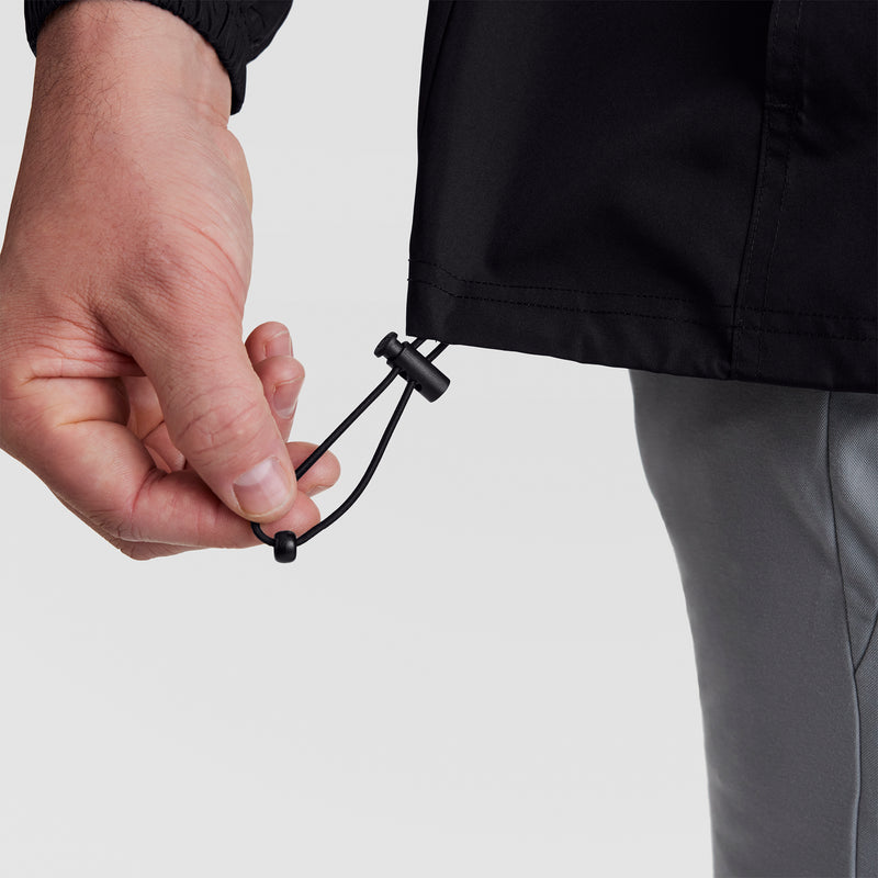A close-up of a hand adjusting the hem drawstring on the black Champion Stadium Packable Jacket, Script Logo - Big & Tall. The wearer has on grey pants against a plain, light background.