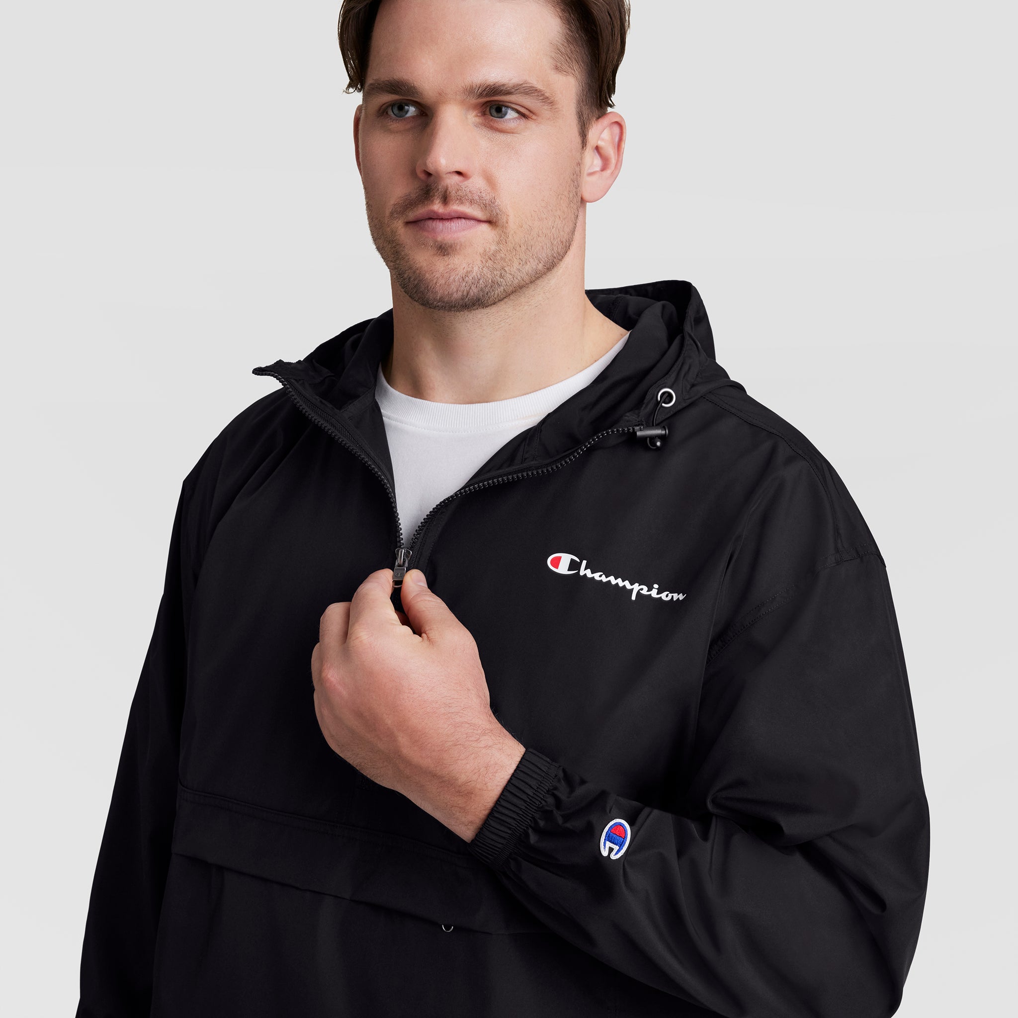 A man in a black Champion Stadium Packable Jacket, Script Logo - Big & Tall, with a white shirt underneath stands against a plain background, zipping up the wind- and water-resistant jacket featuring Champion logos on the chest and sleeve.