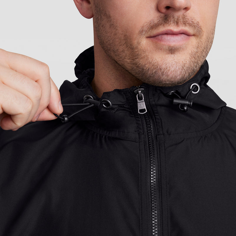 A person adjusts the hood drawstring of a Champion Stadium Packable Jacket, Script Logo - Big & Tall, in black, highlighting its wind and water resistance around the upper chest and face.