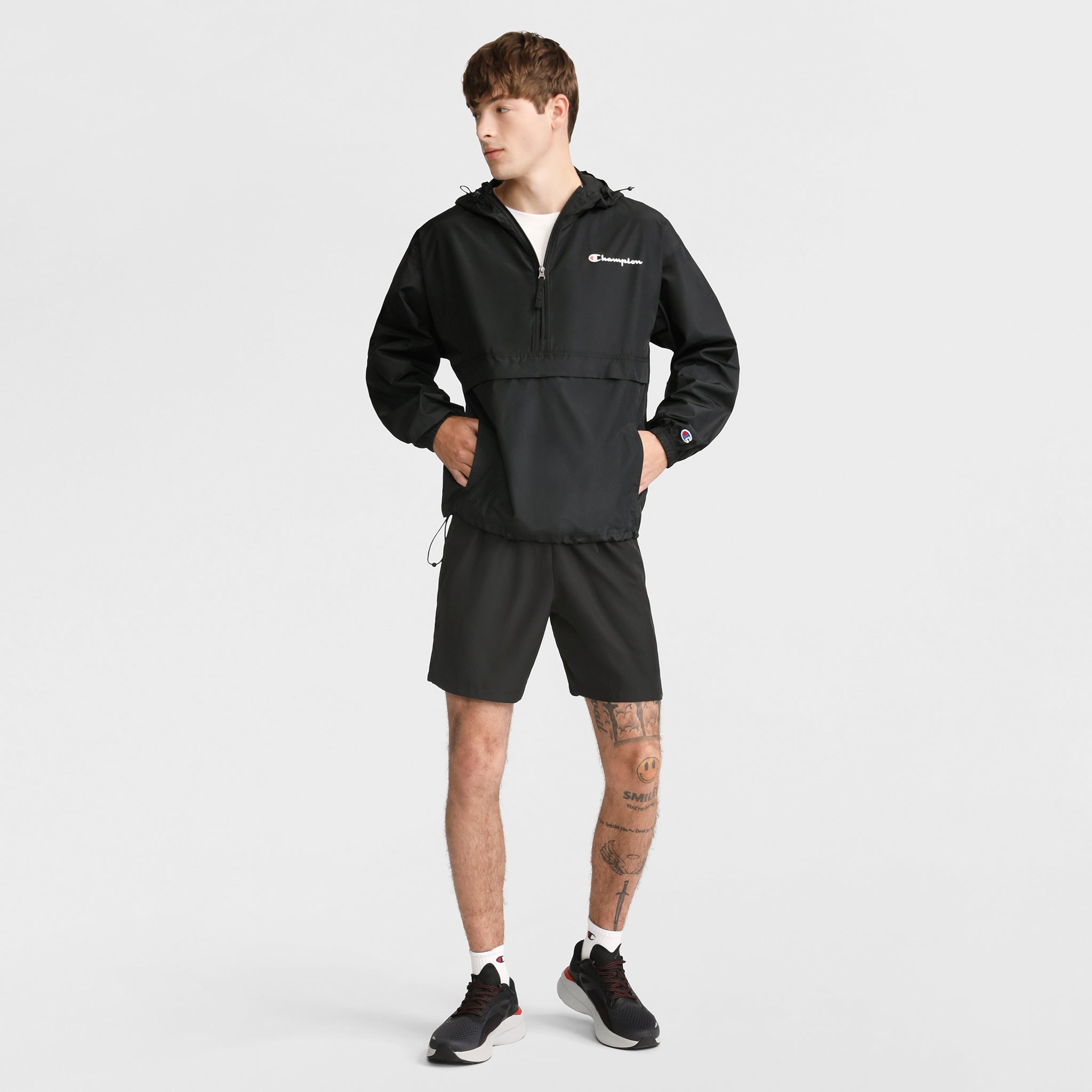 A young man in a black Champion Stadium Packable Jacket with Script Logo, black shorts, white socks, and black sneakers stands against a white background. He has visible leg tattoos and looks to the side with hands in his jacket pockets.
