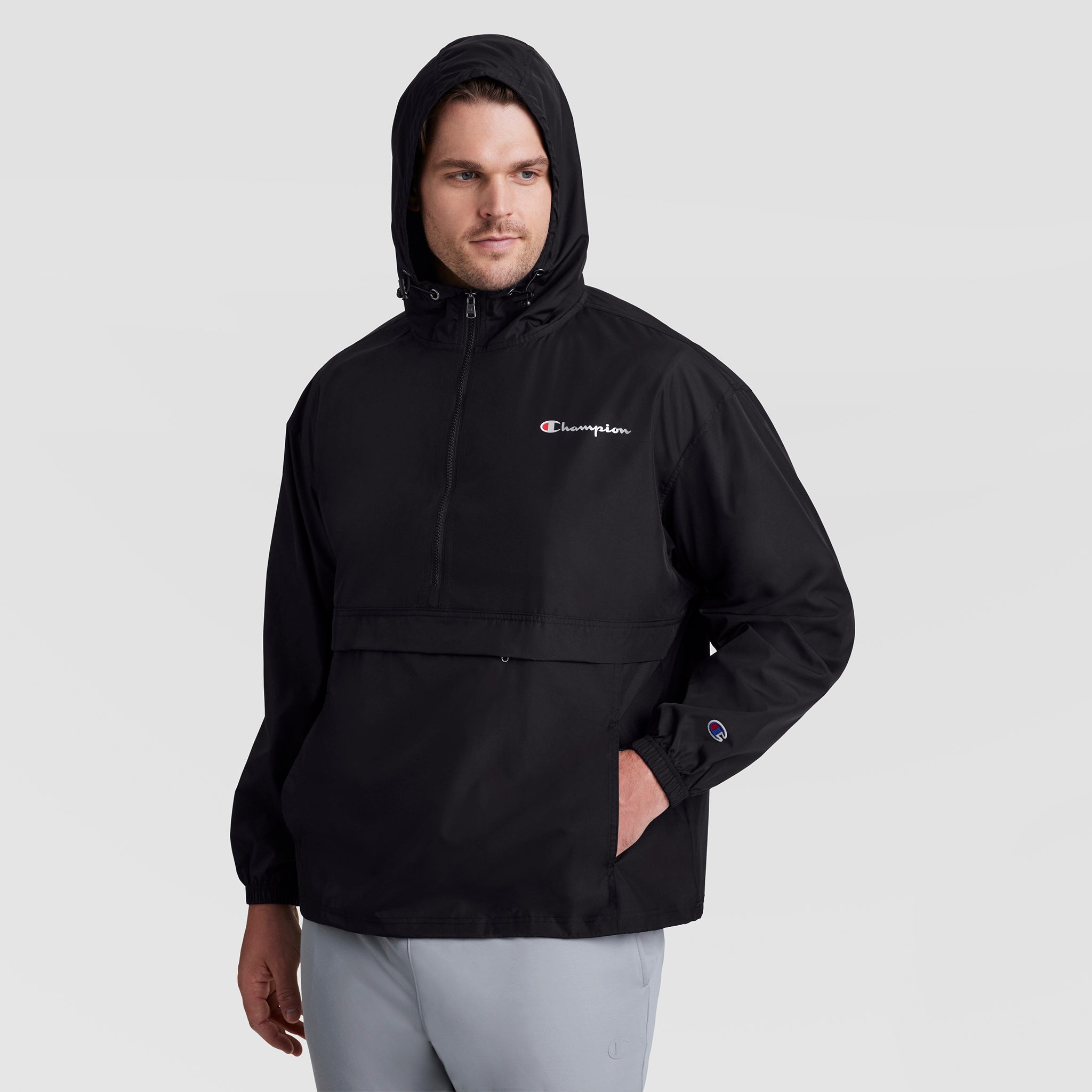 A man wearing a black Champion Stadium Packable Jacket, Script Logo - Big & Tall, with a front zipper and hand pocket, stands against a plain light gray background.