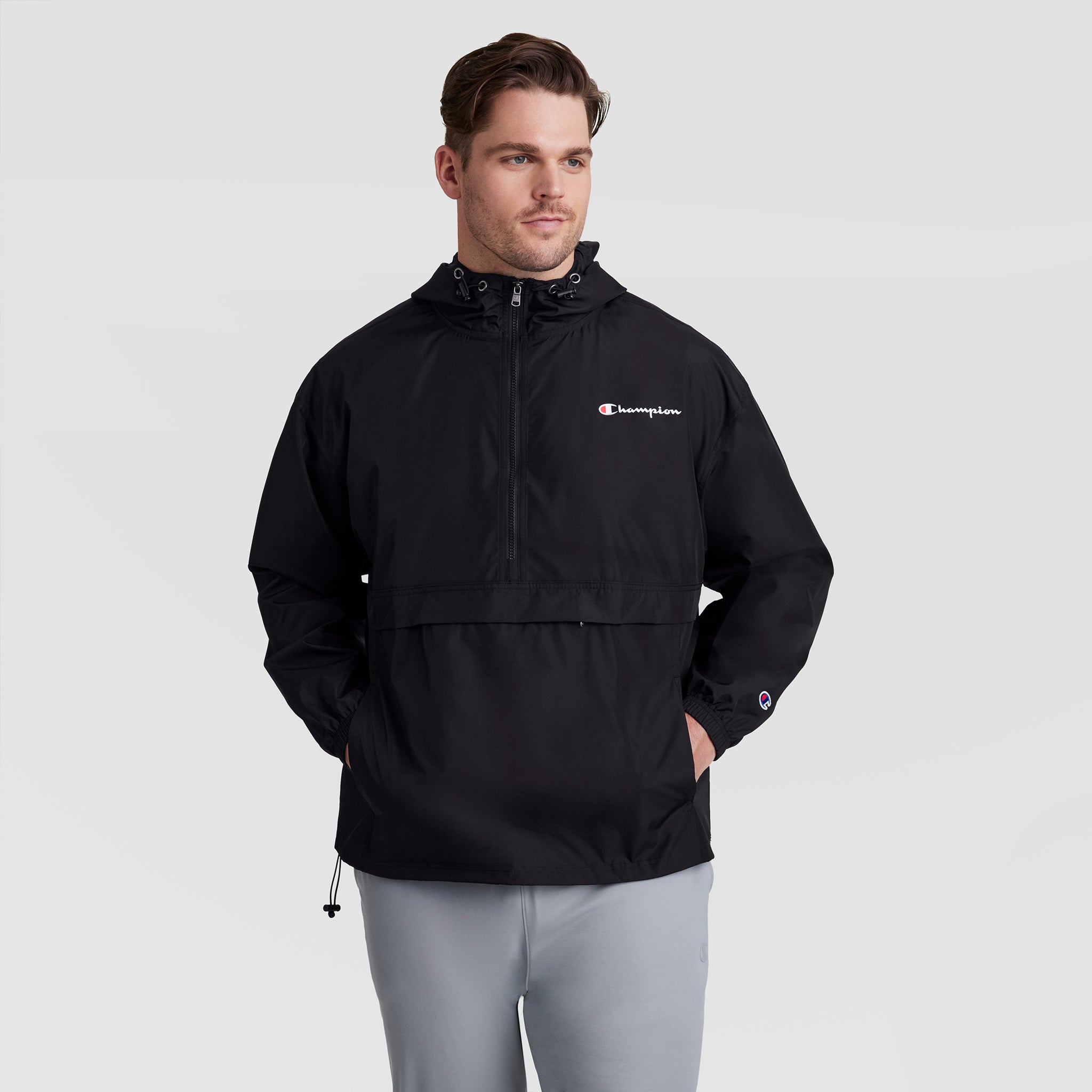 A man with short brown hair stands against a light gray background wearing Champion’s Stadium Packable Jacket, Script Logo - Big & Tall in black, featuring a front pocket and paired with light gray pants. His hands are in his pockets.