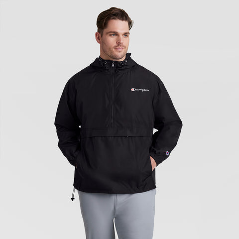 A man with short brown hair stands against a light gray background wearing Champion’s Stadium Packable Jacket, Script Logo - Big & Tall in black, featuring a front pocket and paired with light gray pants. His hands are in his pockets.