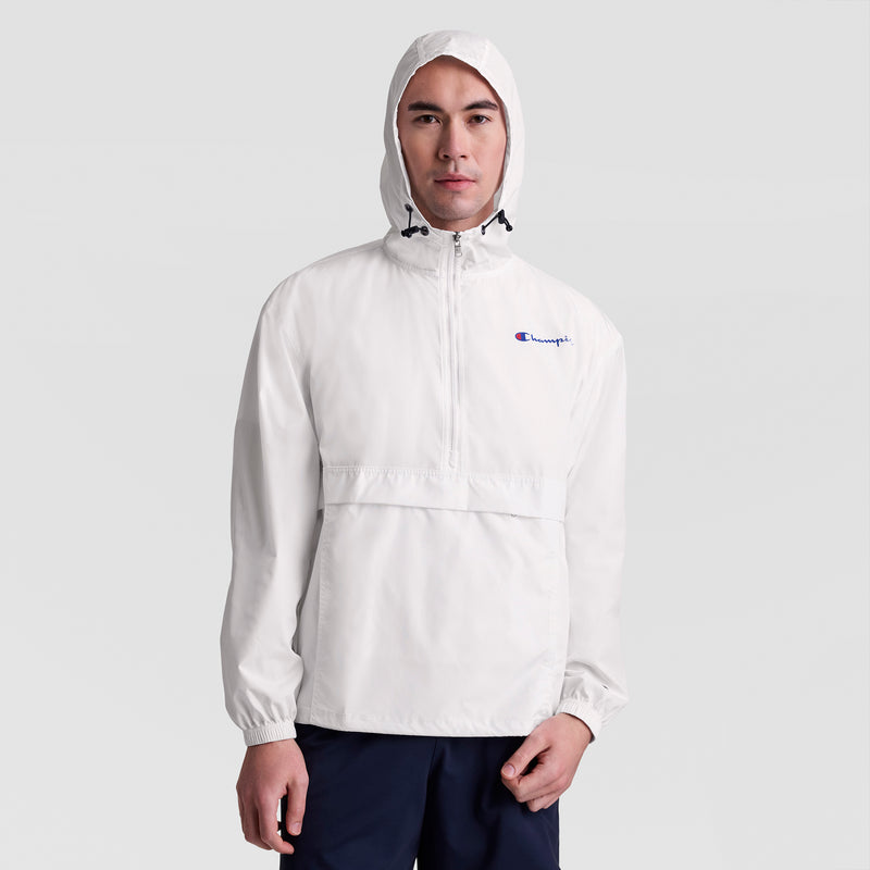 A man wears a white Champion Stadium Packable Jacket with script logo, wind and water-resistant, featuring a front zipper, paired with navy blue pants, standing facing forward against a plain light gray background.