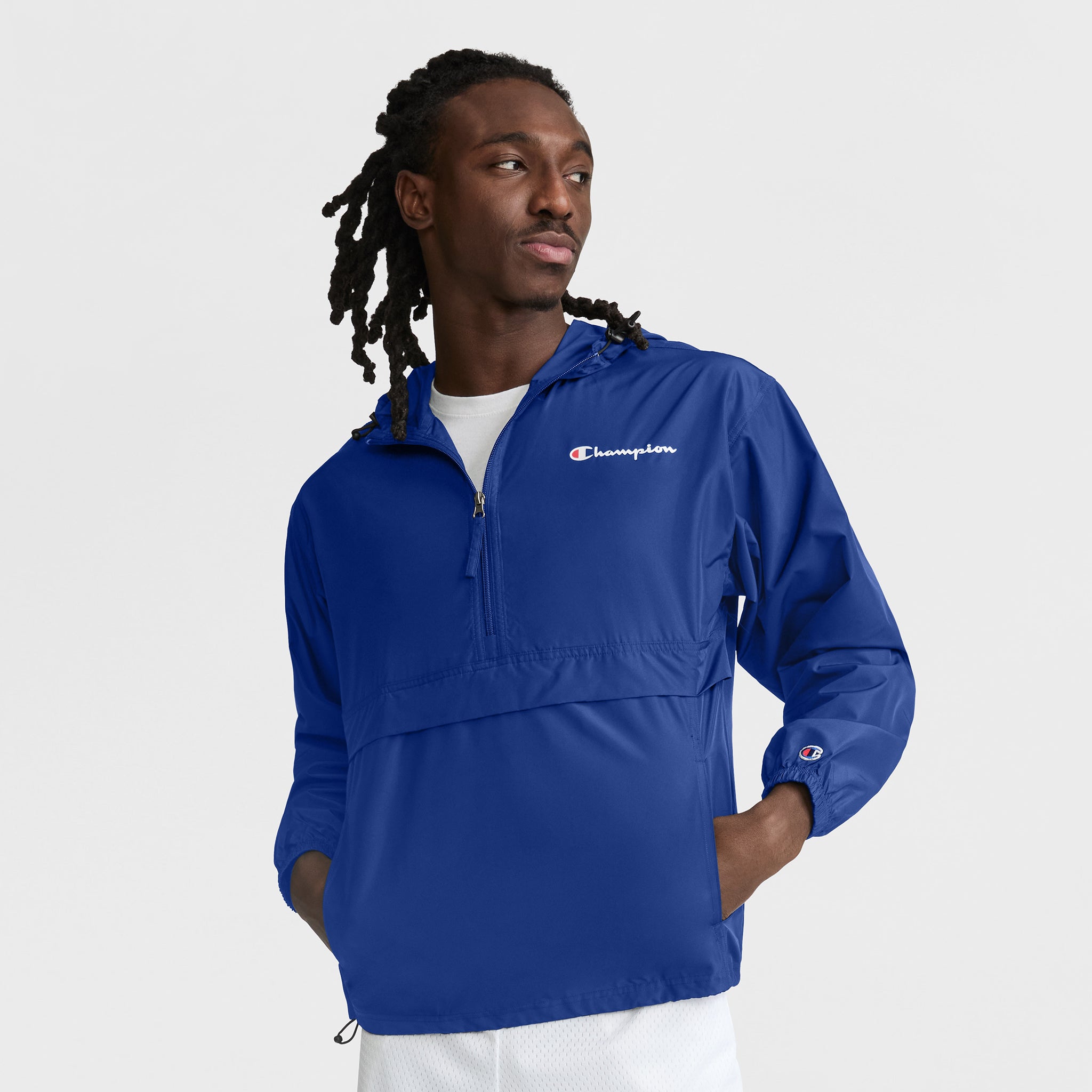 A man with long, braided hair wears the Champion Stadium Packable Jacket, Script Logo in royal blue. The ultra-lightweight unisex jacket features a quarter-zip and front pocket. He stands against a white background, looking left with one hand in his pocket.