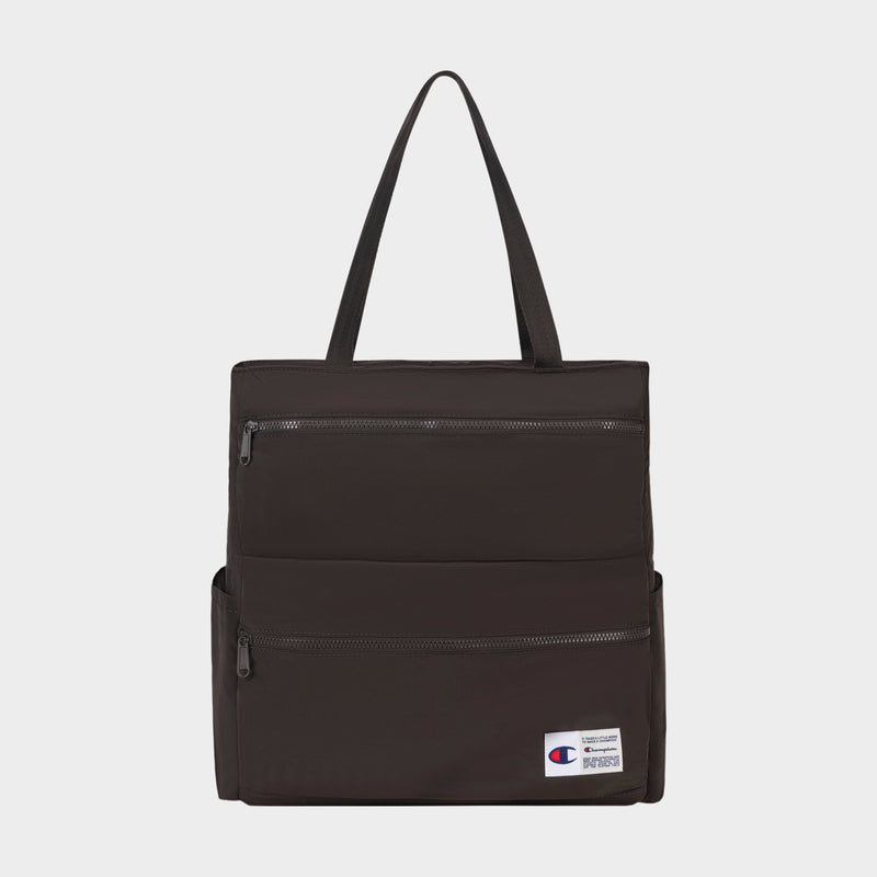 The Champion Explore Tote is a black bag crafted from durable crinkle nylon, with two zippered pockets on the front, side pockets, a padded laptop sleeve, and a small Champion logo patch near the bottom right corner.