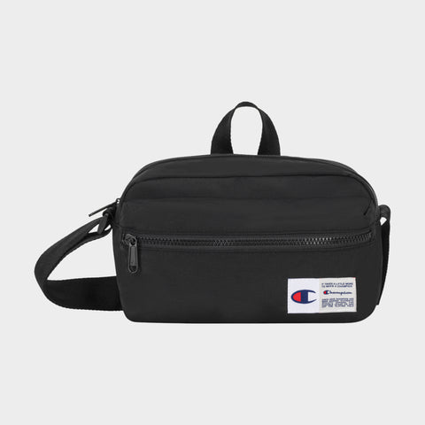 The Explore Crossbody by Champion is a black nylon bag with an adjustable strap, a front zipper pocket, and a small white Champion logo patch on the lower right corner. It is shown against a plain light background.