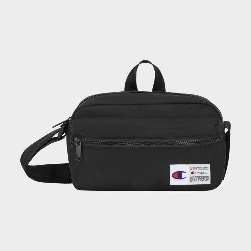 The Explore Crossbody by Champion is a black nylon bag with an adjustable strap, a front zipper pocket, and a small white Champion logo patch on the lower right corner. It is shown against a plain light background.
