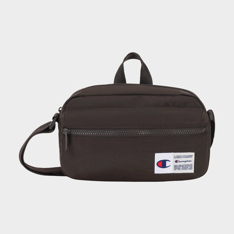 The Champion Explore Crossbody is a small black bag crafted from durable crinkle nylon, featuring a front zipper pocket, top handle, and adjustable strap. A Champion logo patch accents the lower right corner. The product is displayed against a plain white background.