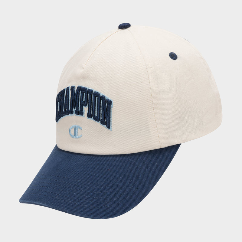The Amor Adjustable Baseball Cap by Champion features beige cotton twill, “CHAMPION” embroidered in dark blue on the front, a small light blue C logo below, and a navy blue curved brim.