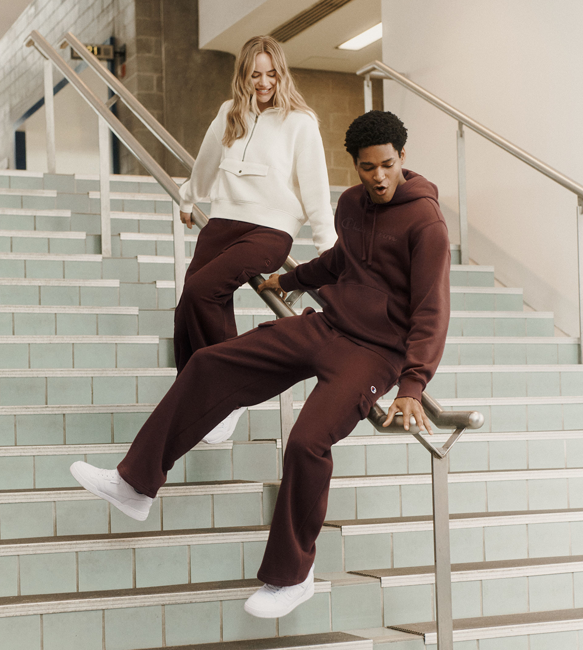 A man and a woman wearing matching burgundy sweatshirt and jogger sets pose together on a tiled indoor staircase.