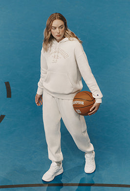 A woman stands on a blue basketball court wearing a matching off-white/cream hoodie and jogger sweatpants set, holding a basketball.