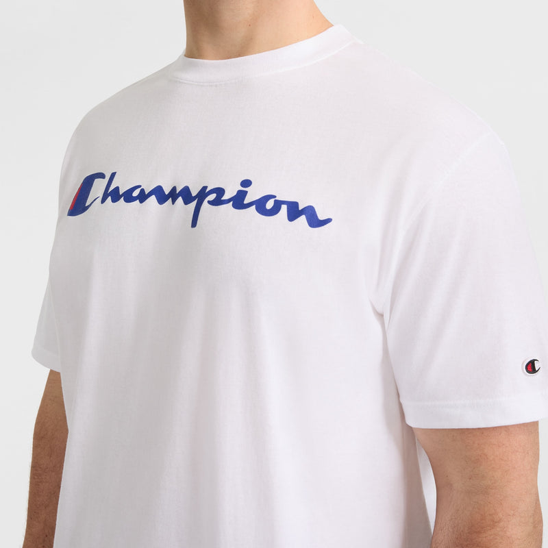A man wears the Champion Classic Graphic T-Shirt, Script Logo - Big & Tall in white, featuring a bold blue logo across the chest and a small sleeve emblem—ideal for casual streetwear.